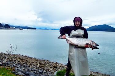 A person in a beanie and plastic apron holds a large shiny silver fish on a rocky bank with a wide river behind her.