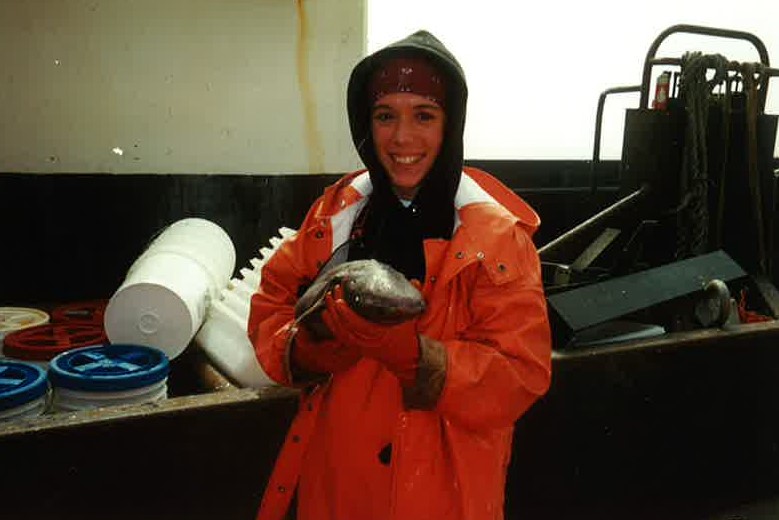 Image of smiling woman in orange rain gear cradling a baby shark with buckets and laboratory equipment in the background.
