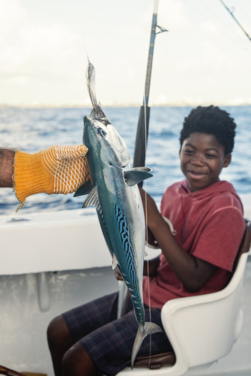 dam-migration-miss/md_jIMIsdZFYG91.jpg Young black boy sits on a white chair holding a fishing pole and smiling. Yellow gloved hand comes into foreground from the left holding a fish upright.