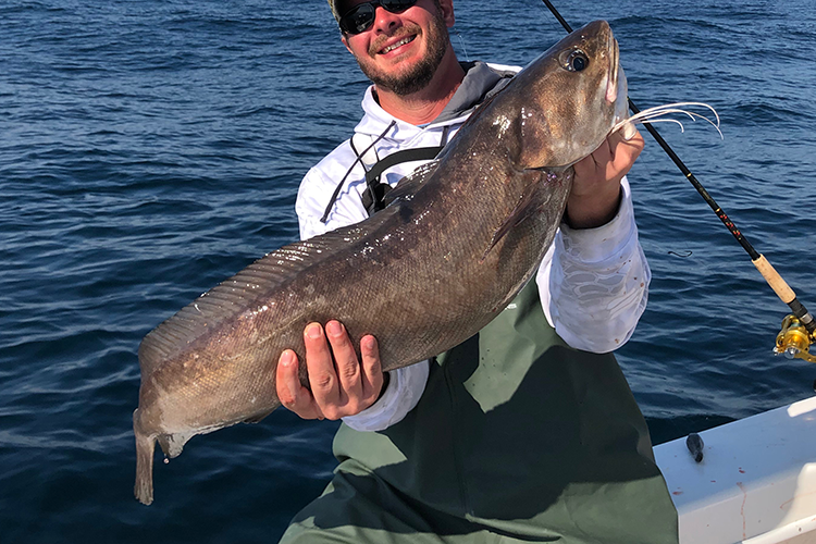 Causasian male with sunglasses sits on the edge of a boat holding a white hake in both hands.