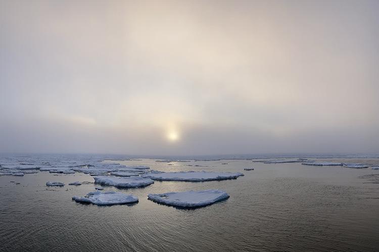 Sun shining through fog over water and sea ice.