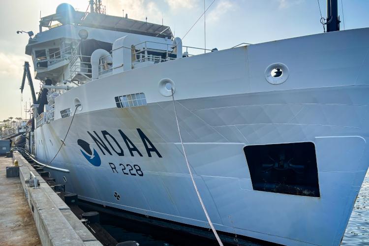 A large white ship with the NOAA circle emblem is at dock.