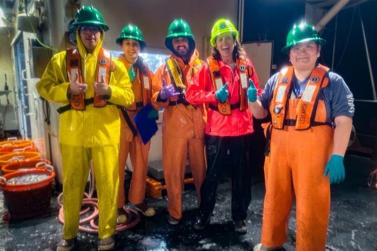 5 men and women wearing hard hats, gloves, and life vests stand smiling on the deck of a ship at night. 