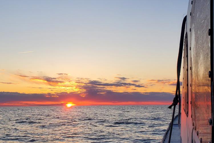 Sun setting just above the horizon. There’s a thin layer of clouds just above the horizon and the color of the sky ranges from light blue to peach to orange with a touch of purple. The photo was taken from the side of a commercial fishing vessel and part of the vessel’s side is in the photo.