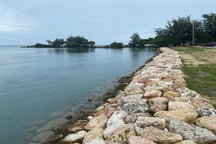 Under a grey sky, calm waters lap a coastal revetment project that extends along a sandy, grassy shoreline, with trees popping up in the distance on a very small peninsula and on the edge of the island.