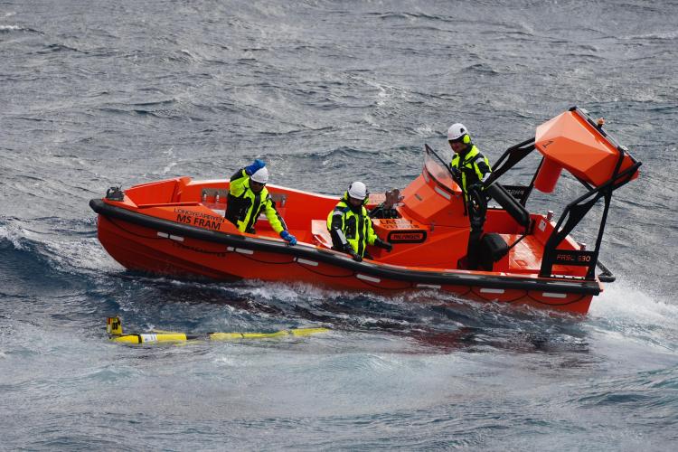 A small orange boat pulls up alongside a yellow glider floating at the ocean surface. There are three people in black and yellow safety clothing on the boat.