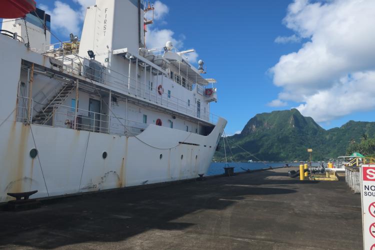 NOAA Ship Hi'ialakai is tied up at a fuel pier in Tutuila with a green, forested mountain in the background.