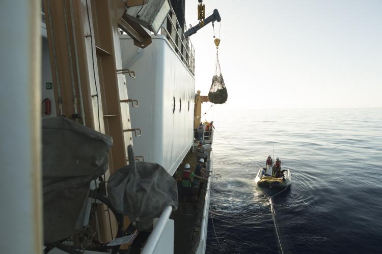 A crane off a big NOAA ship picks up a large bag of marine debris off a small boat below. 