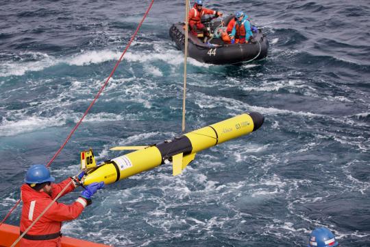 A bright yellow glider being lowered into the ocean. scientist in Zodiac assisting with deployment.