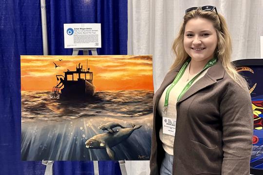 Megan Amico stands next to her painting at the World Fisheries Congress 2024 conference. The painting is an under and above water scene. Above the water, the sky is bright orange with some yellow clouds. There is a commercial lobster boat hauling up a lobster pot. Below the water is a North Atlantic right whale mother calf pair swimming under the boat.