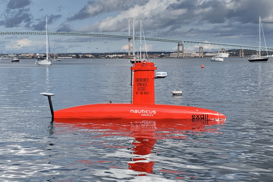 A small vessel shaped like a submarine floats in calm ocean waters, with a large suspension bridge in and sailboats in the background.