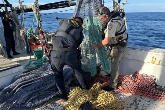 Two men in vests and khakis stand on a commercial fishing boat with nets at their feet. They are inspecting a net hanging between them.