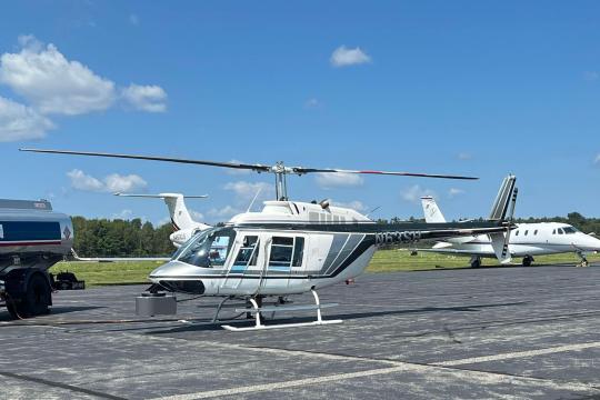 A mostly white helicopter in the center of the image on an airport runway. Small white passenger aircraft are parked in the background and there is a fuel truck to the left.