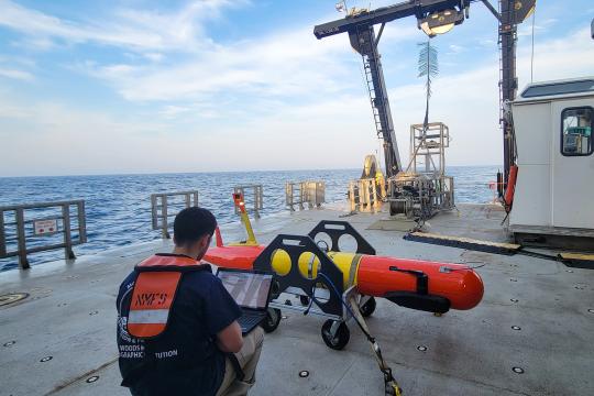 A scientist wearing a personal flotation device squats in front of a long-range autonomous underwater vehicle typing on a laptop while on the back deck of a research vessel at sea.