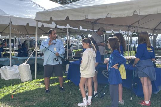 A white man in a blue button down shirt and shorts standing next to a tent holds a fishing reel while several children watch.