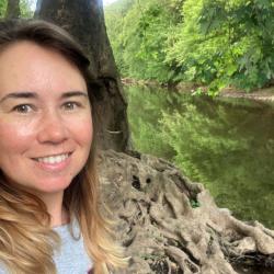 Selfie of woman smiling standing next to a tree along a riverbank. 