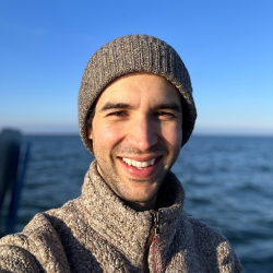 Brian Galvez in sweater and cap by the water.