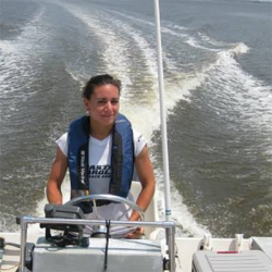 Women with blue flotation device driving a small craft, wake in the background.
