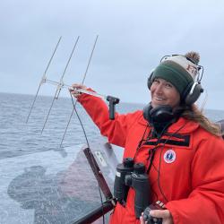 Heather Foley in orange jacke, binoculars and antenna in hand on board boat. 