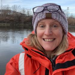 Female scientist wearing orange jacket and blue hat smiling and standing in front of a wetland.