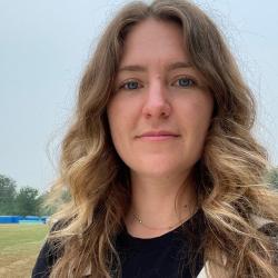 Female scientist wearing white overalls and a black t-shirt in front of a field.
