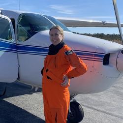 Women in orange flight suit standing in front of small plane. 