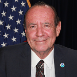 A White man wearing a suit and tie and a NOAA pin smiles in front of an American flag and black background.
