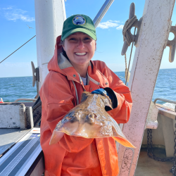 Smiling woman wearing orange survival suit holds a fish on a boat.