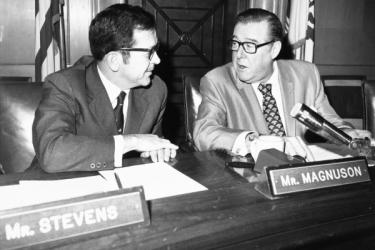 U.S. Senators Ted Stevens (R-AK) and Warren Magnuson (D-WA) having a conversation in their neighboring senate seats, with state flags in the background.