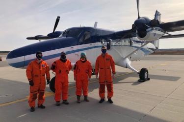 Two NOAA pilots and two survey scientists dressed in orange jumpsuits stand in front of the NOAA Twin  otter airplane.