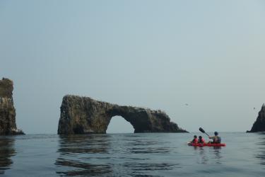 Three people in a red kayak paddle near a large stone arch.
