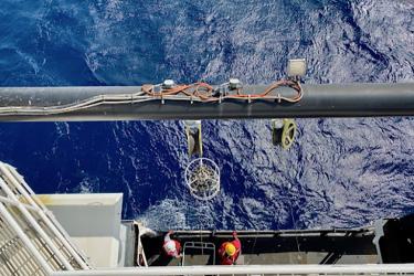  A white circular frame with 12 gray bottles is suspended over the ocean water from an A-frame on the side of the ship. The image is taken from above the A-frame. It is daytime in good weather. Two crew in hard hats stand on the deck near the rail, beneath the sampling gear.