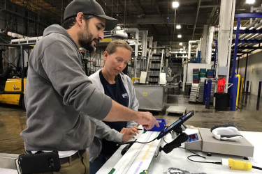 In a large open space in an industrial building, two people are using an electronic measuring board on a tabletop. The measuring board is long and narrow. A digital scale is also on the table. Both are connected to an electronic table that collects and records the lengths and weights of squid laid on the board.