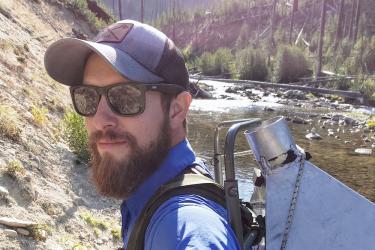 Smiling male wearing sunglasses and cap is carrying a backpack while hiking near a river.