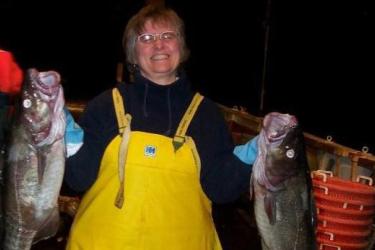 Women in foul weather gear overalls holds up two cod. In the background are baskets and fish on the deck for sorting from a bottom trawl