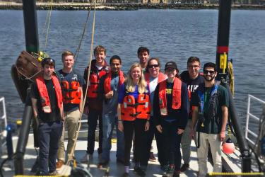 Ten people wearing orange life vests stand at the back end of a research vessel with a large, metal, black arch above them. The vessel is on the water with several buildings on the shore in the background. 