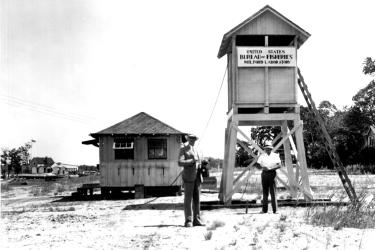 Two men standing in front of an elevated shack with a Bureau of Fisheries sign and a ladder alongside. A second building can be seen in the background. 