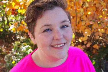 Kristin is a white person looking up at the camera smiling with short reddish brown hair wearing a bright pink sweater with a background of yellow leafy bushes and green foliage at the United States Botanical Garden.