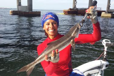 Woman in red shirt holding caught cobia, with water in the background.