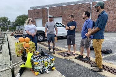 A color image taken in the summer during the day in overcast conditions. Two wire cages containing on-demand fishing gear are at left at the edge of the docking area that abuts the cement parking lot and lay-down area. The group is at the center. A brick building with two garage bays is in the background.