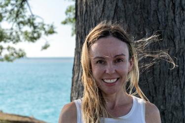 Portrait of a woman smiling in front of a tree with the blue ocean in the background.