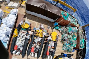 An shot from above, looking down on the deck of a ship, showing 4 small boats, many team members, and large piles of derelict fishing gear.