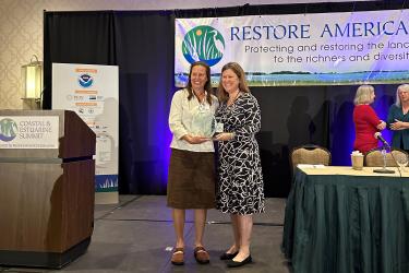 Ciona Ulbrich receiving a glass trophy from Carrie Robinson on stage at the Restore America's Estuaries Coastal & Estuarine Summit in New Orleans, Louisiana