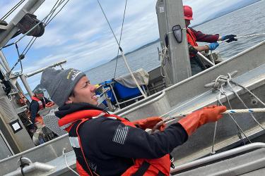 In the foreground, a person wearing a black hoodie, red life jacket, and red rubber gloves stands on a ship and pulls on a rope. Two additional people are seen in the background, also wearing red life jackets.