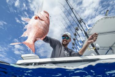 a man on a boat in sunglasses and a hat holds a caught redfish on his line
