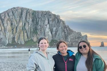 Kaitlin Palmer, Kourtney Burger and Cory Hom-Weaver stand in front of Morro Bay rock during sunset. The rock is a prominent feature in Morro Bay that jets up from the sand and ocean over 500 feet tall. 