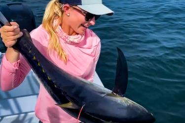 A woman in a hat and glasses on the deck of a boat holds a tuna with a red, string-like object attached near its top fin