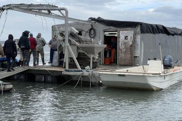 A metal floating wharf with a small boat attached. A small group of people standing on the wharf are talking. A metal machine for processing oysters is on the wharf, filled with oysters. 