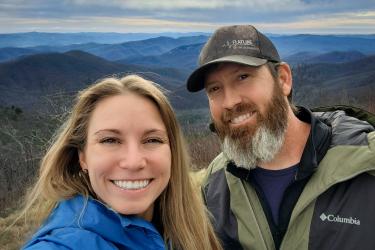 Two people smiling in front of a vista of a blue-tinted mountain range