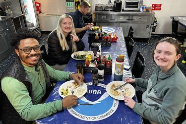 Three people face the camera at a dinner table with a NOAA logo table cloth. Each person has a plate of food in front of them on the table and a smile on their face.
